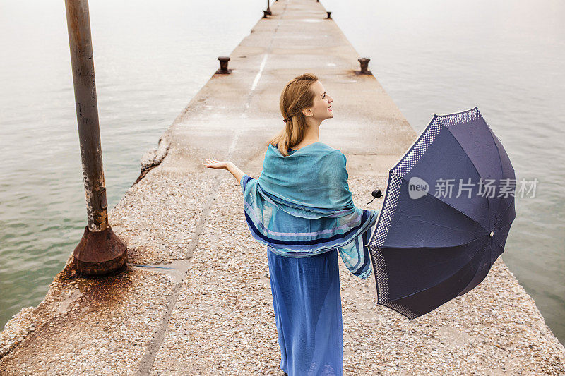 下雨天，穿蓝衣服的女人撑着伞站在码头上