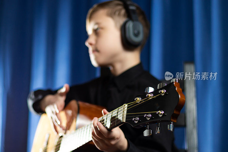 一名13岁的少年穿着黑色衬衫，头戴耳机，拿着吉他，在音乐工作室的蓝色背景下录制了一段旋律。有选择性的重点。