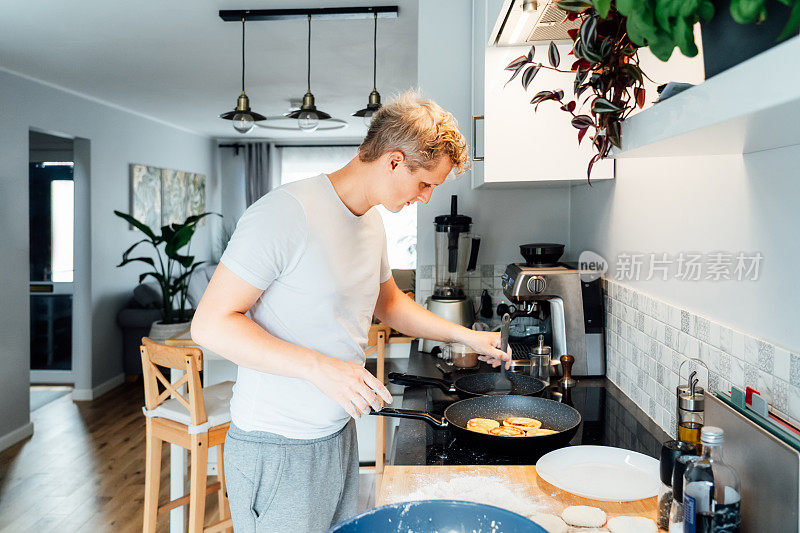 一个年轻人在家里现代化的轻厨房里做起司煎饼。一名男子正在准备在平底锅上煎的煎饼。家务和家务。烹饪的爱好。有选择性的重点