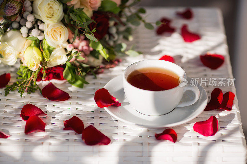 一杯茶，玫瑰花瓣，金婚戒指和一束美丽的花束放在白色的桌子上。婚礼庆典的概念。新娘的早安。