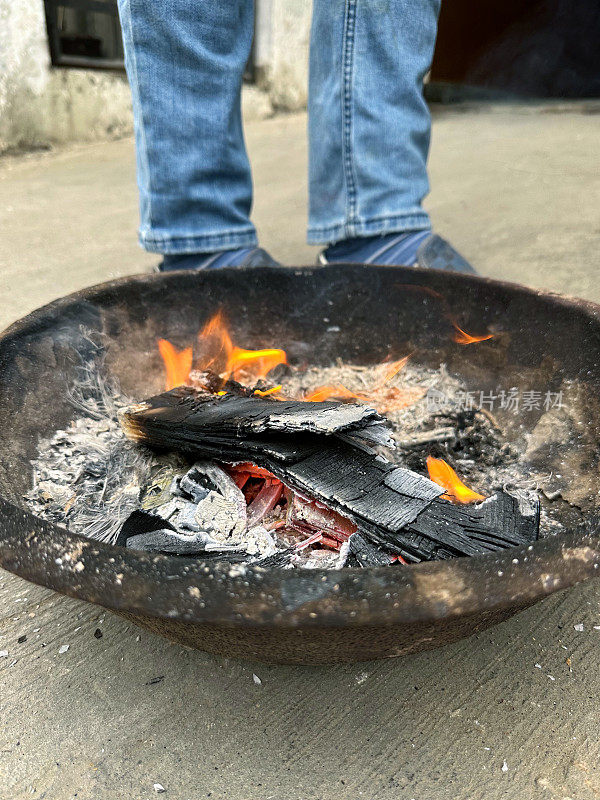 卡代(印度火碗)的特写图像，舞蹈的火焰，灰烬和烟雾，燃烧的木头余烬，无法辨认的人在取暖，高视图，关注前景