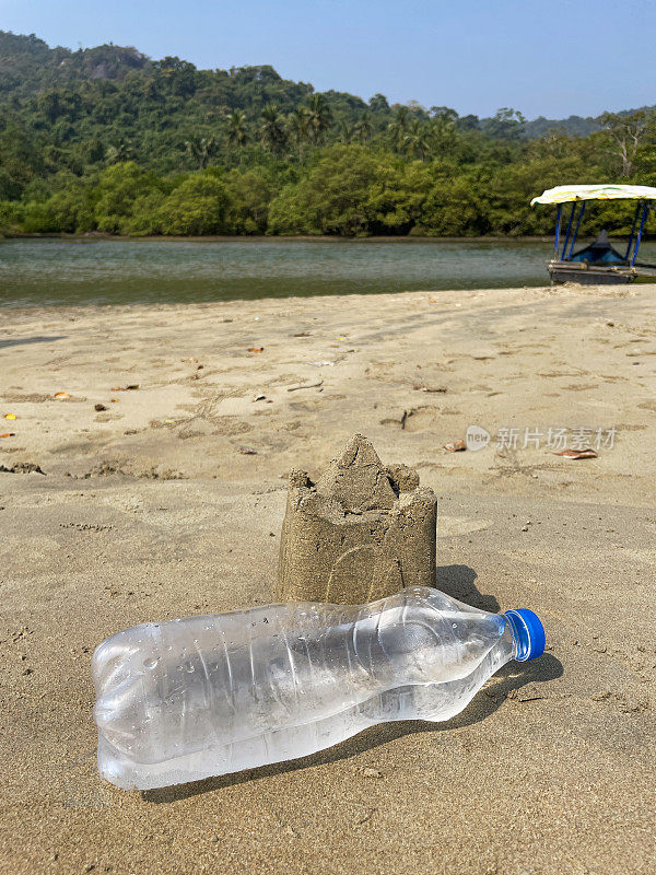 干地上用水桶和铁锹制作的沙堡外的塑料水瓶特写，沙滩，海岸上的海洋垃圾和污染，乱扔的沙子，渔船，有棕榈树的林地，重点在前景