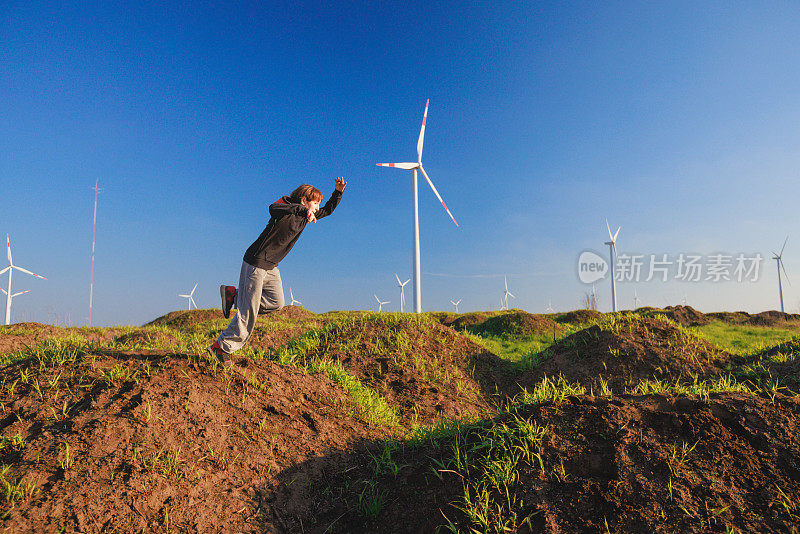 男孩在乡村的绿色山丘上奔跑和跳跃，在风力涡轮机旁玩耍