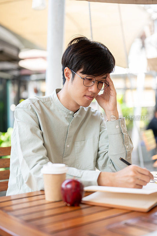 一名专注的亚洲男子坐在咖啡馆的户外桌子旁，把自己的想法记在一本书上。