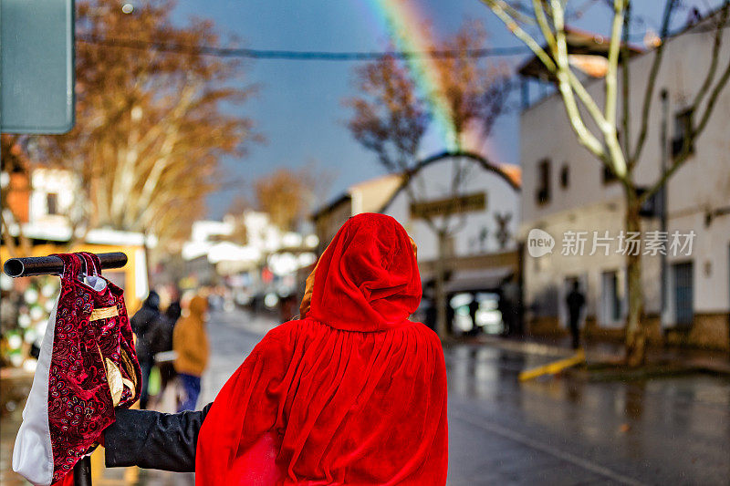 一个穿着红色丝绒长袍、戴着兜帽的女人的后视图，在一个下雨的下午，在被雨水浸透的村庄街道上，看着彩虹。