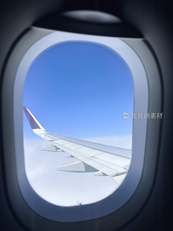 飞机机舱窗户的图像，透过飞机机翼的窗户看到云层上方，飞过蓬松的白云，小翼和板条，蓝天，聚焦于前景