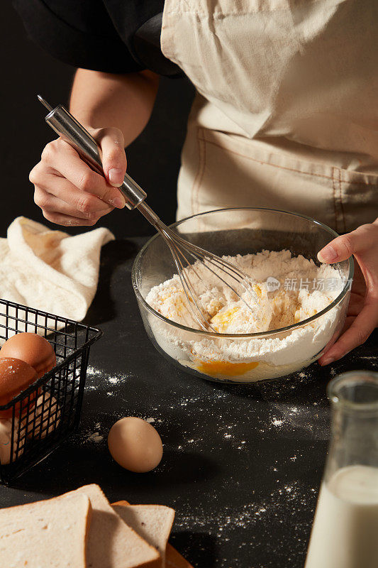 戴着围裙的面包师正在玻璃碗里用打蛋器搅拌鸡蛋和面粉。黑色的桌子上放着一篮子鸡蛋、三明治和一瓶牛奶