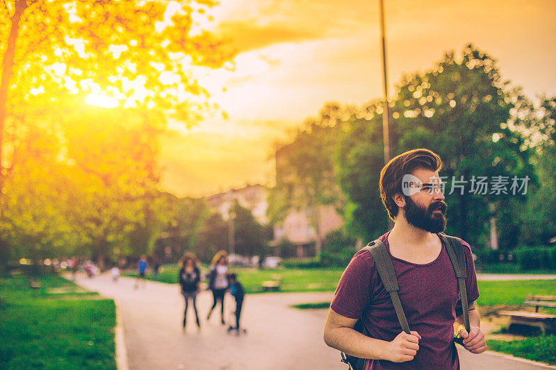 在一个阳光明媚的夏日，一个背着背包的帅气胡子男人在公园里