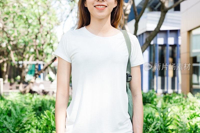 亚洲嬉皮女孩棕色长发在白色空白t恤站在街道中央。一位身着街头服饰的女性站在绿色的城市背景上。为文本或设计留出模拟空间。