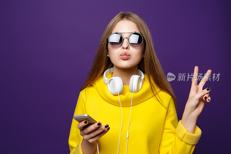 描绘年轻的少女戴着耳机和电话，穿着黄色毛衣，孤立在紫色的背景上。