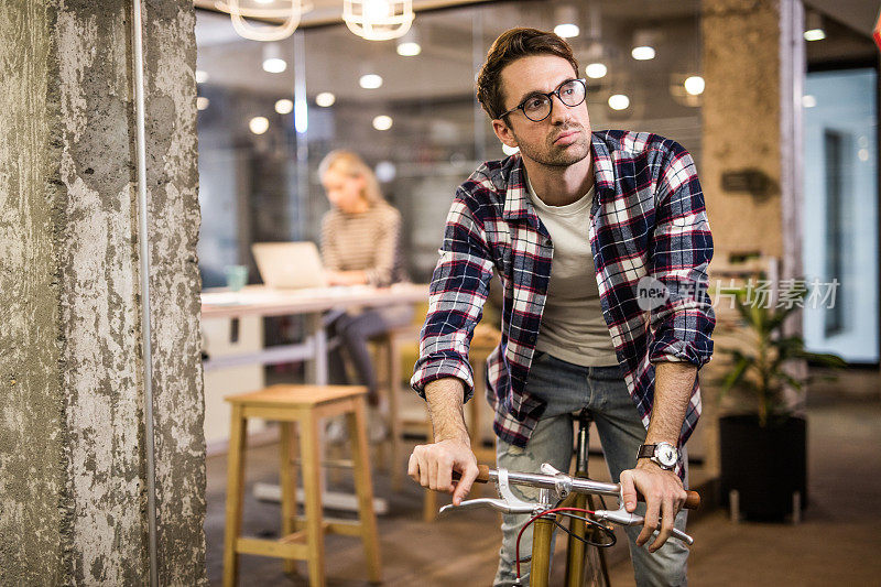 年轻的沉思企业家骑着自行车在一个休闲的办公室。