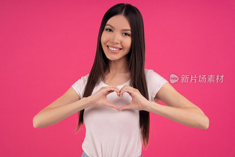 有魅力的亚洲女性做出心形手势，表达爱意，说做我的情人，正面微笑，穿着白色的衣服，对着粉红色的空墙摆姿势。肢体语言的概念