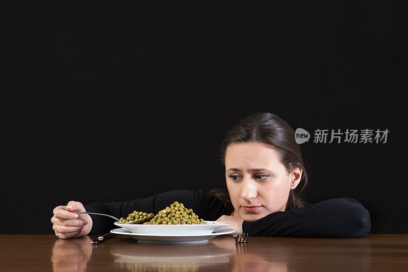 节食。一个不高兴的年轻女人拿着满勺绿豌豆坐在桌子上，盘子上有一顿大的减肥餐