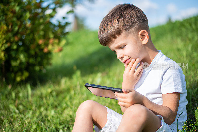 夏天日落时，兴高采烈的孩子坐在草地上，看起来就像手机里的卡通。可爱的男孩在大自然中玩耍