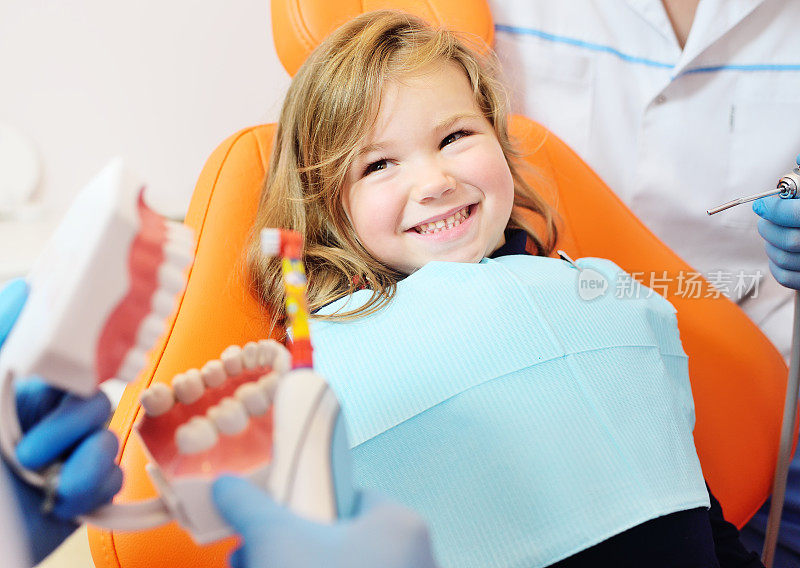 儿科牙医向坐在橙色牙科椅上微笑的小女孩展示如何正确刷牙
