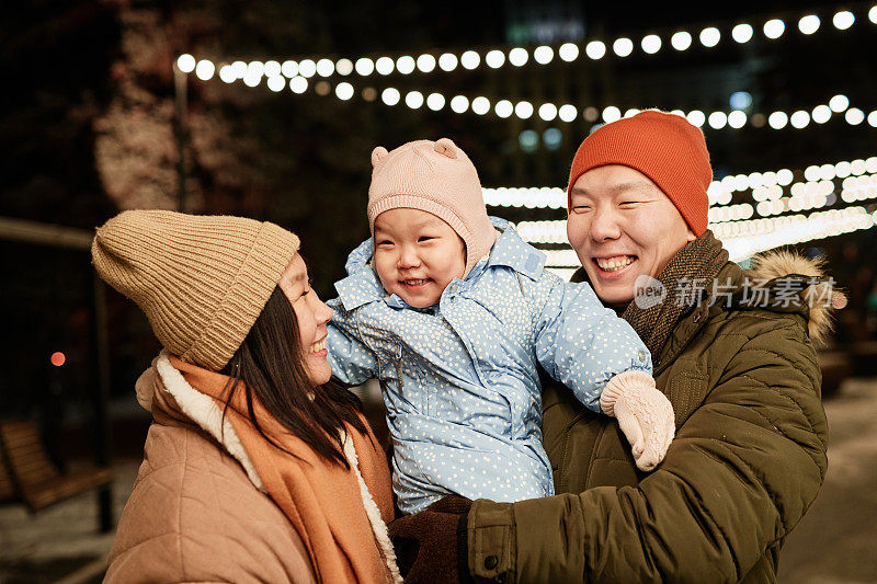 水平中景拍摄的快乐的亚洲夫妇和他们可爱的蹒跚学步的女儿在户外度过的冬季夜晚