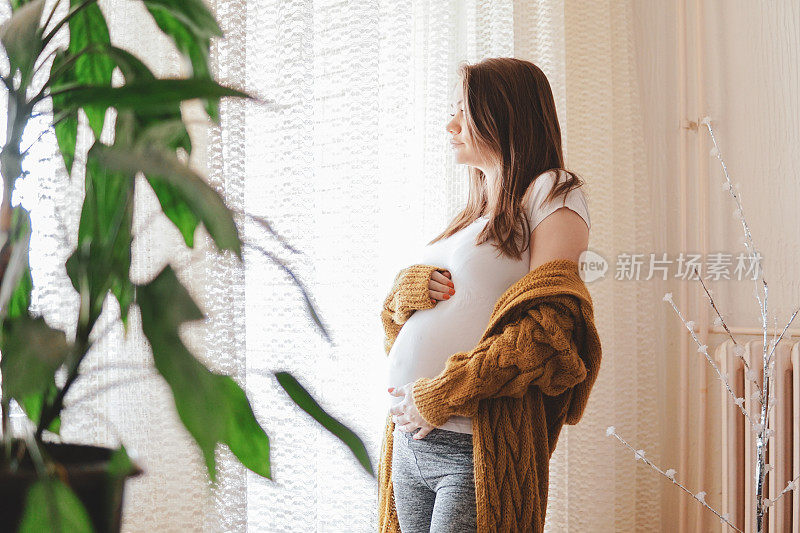 幸福的孕妇站在窗边，抱着她的肚子