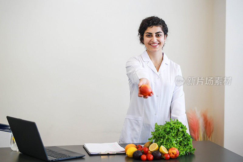 年轻漂亮的女性健康营养师站在她的桌子后面，拿着苹果，健康的蔬菜在桌子上健康的饮食