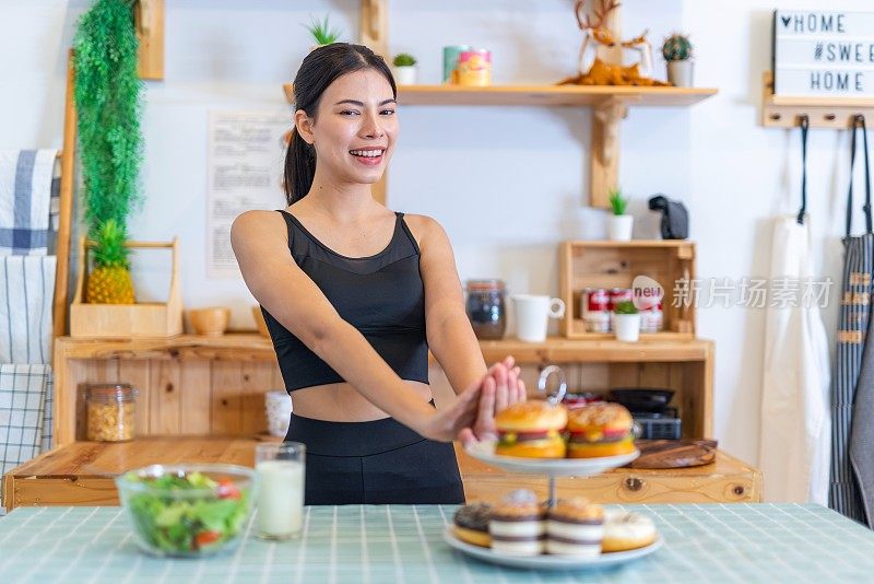 照片年轻美丽的亚洲女性女士在运动服装说不和拒绝诱惑汉堡和甜甜圈在厨房的桌子上，选择一碗沙拉与樱桃番茄和一杯牛奶的饮食和减肥健康的生活方式