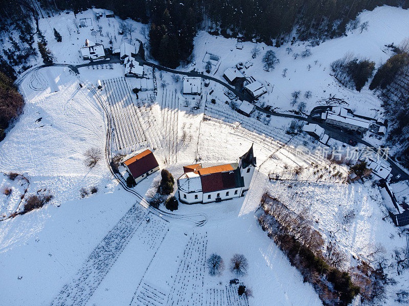 鸟瞰图:一座孤零零的教堂坐落在一座白雪覆盖的小山上，背景是森林覆盖的山脉。