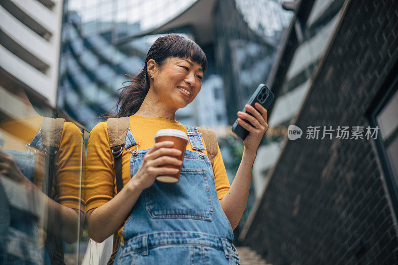 一名日本女子拿着智能手机和一杯咖啡，看着手机屏幕，背景是一栋办公楼