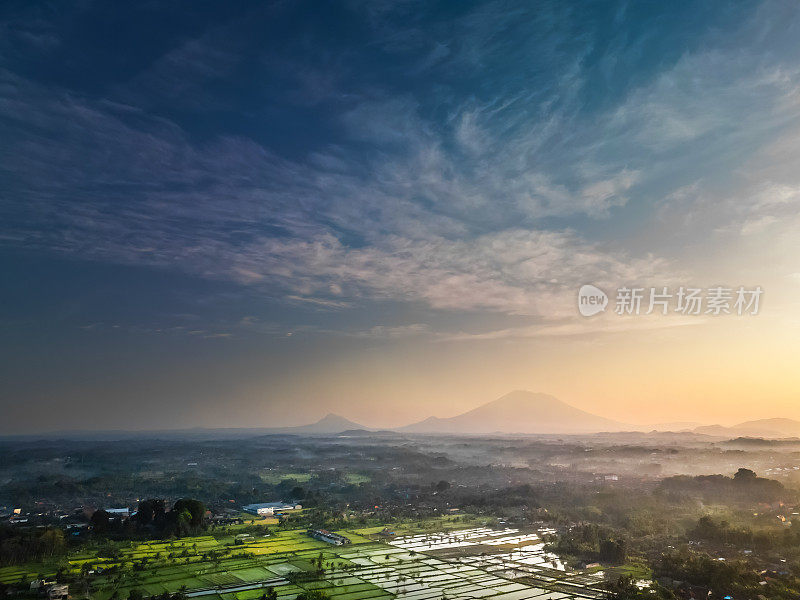 从远处鸟瞰印尼巴厘岛吉安雅的阿贡山，前方是绿色的稻田。巴厘岛乡村风光，稻田梯田花园中的一个村庄，而日出。印尼旅游理念