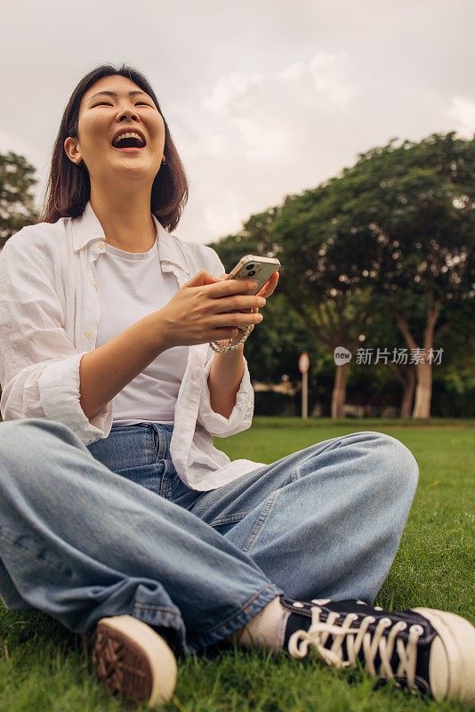 快乐的亚洲女性在城市公园放松时在户外用智能手机发信息