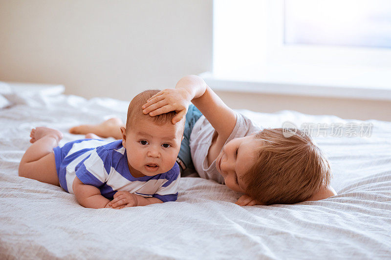 可爱的男孩和婴儿放松和在卧室里玩。小弟弟抱着她刚出生的婴儿。蹒跚学步的孩子认识新兄弟姐妹。