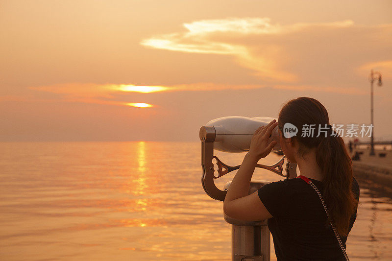 年轻女子透过投币双筒望远镜眺望海洋漫步海上日落