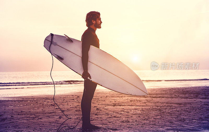 男子冲浪者携带他的冲浪板在日出-潮男穿着潜水衣等待海滩上的巨浪-极限运动概念-聚焦剪影-哑光滤镜与柔软的蓝色vignette