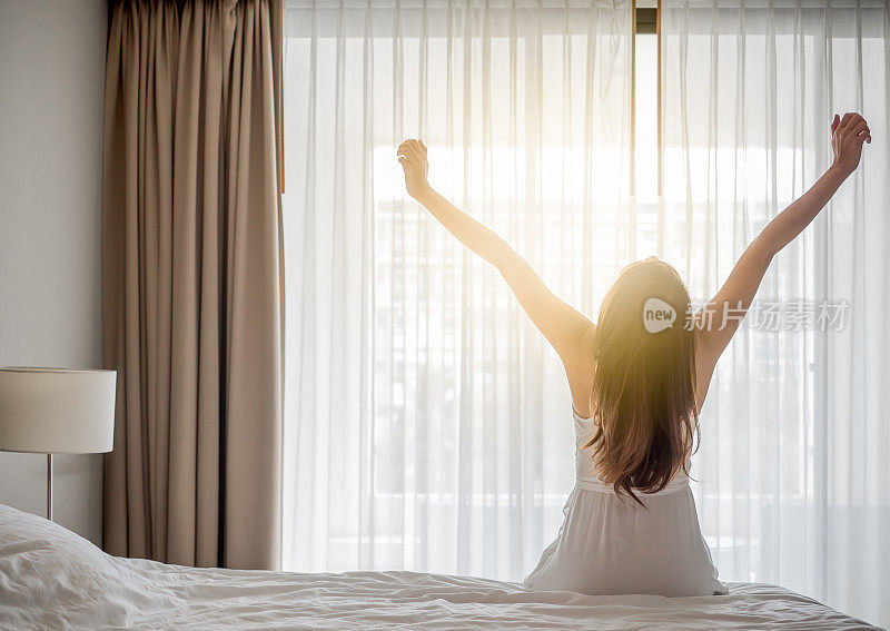 亚洲女人早上醒来，坐在白色的床上伸展，感觉快乐和新鲜