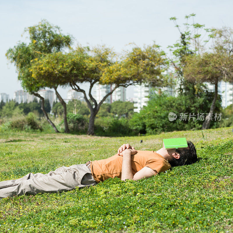 一个成年人躺在绿色的草地上，用书盖住他的脸