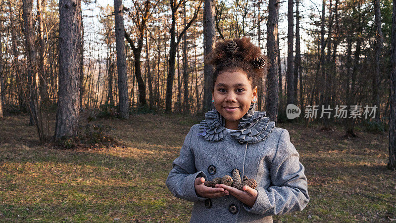 全世界的孩子们。一个快乐的美国黑人女孩在公园里玩松果的肖像。微笑着看着镜头，头发上戴着松果。