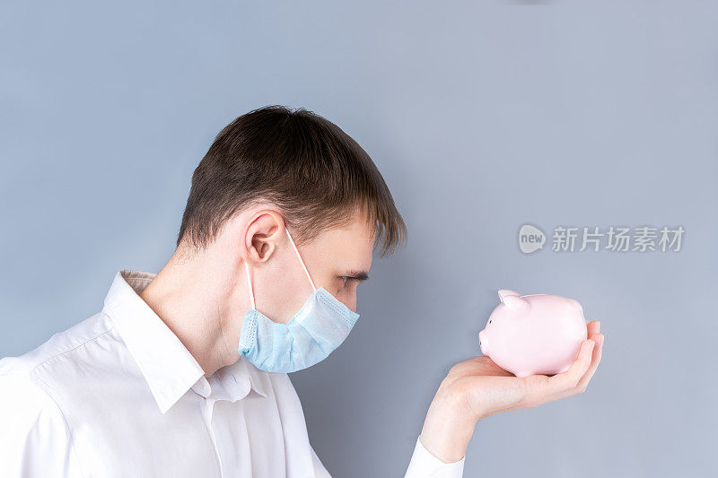 一个手里拿着粉色储蓄罐的人戴着医用口罩，身穿白色衬衫，身穿灰色背景制服。冠状病毒