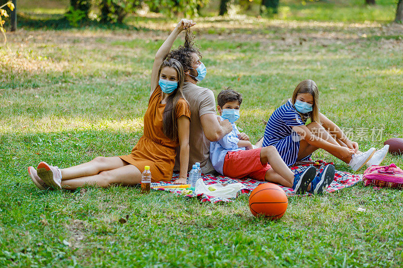 一个戴着防护口罩的家庭正在享受在一个草地野炊在病毒时期。