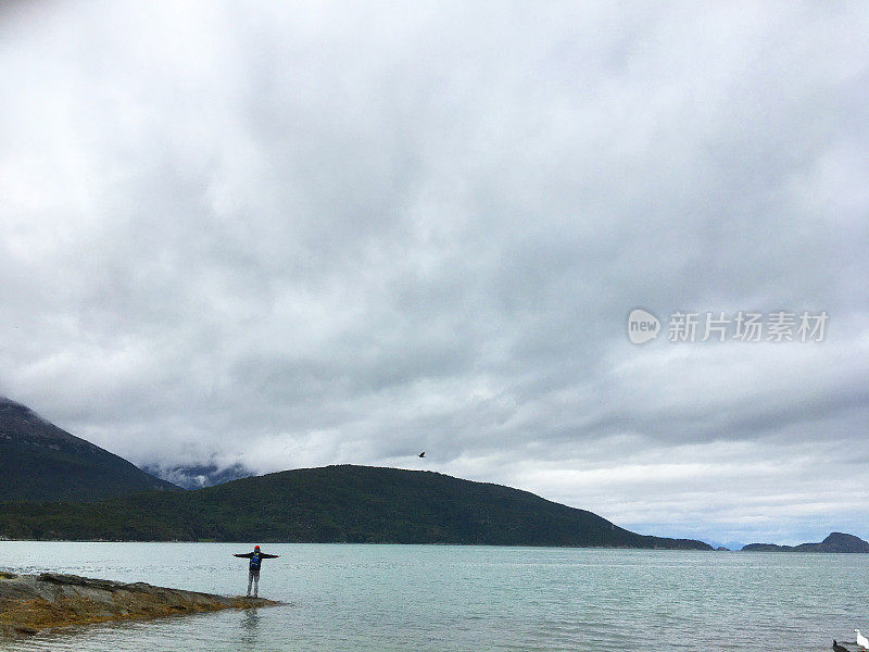 一个多云的日子，在靠近智利边境的阿根廷火地岛省，一名拉丁背包客男性在巴塔哥尼亚绿松石湖前张开双臂。