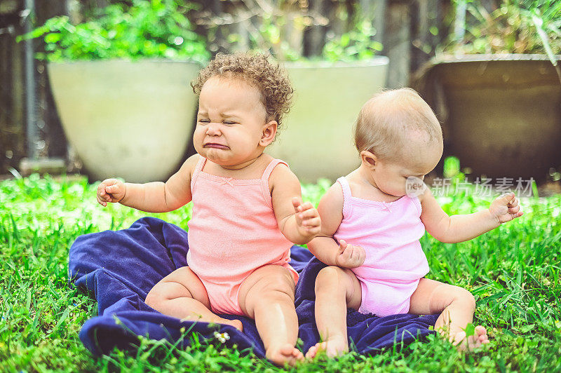 两个婴儿一起坐在外面，他们穿着相同的衣服，年龄相同，但大小不同，种族不同，其中一个在哭