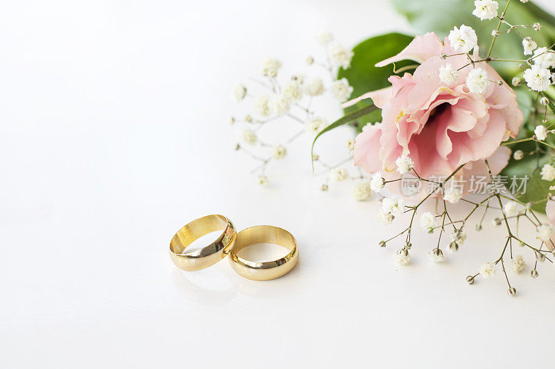 白色背景上有粉红色的花和两枚金色的结婚戒指。