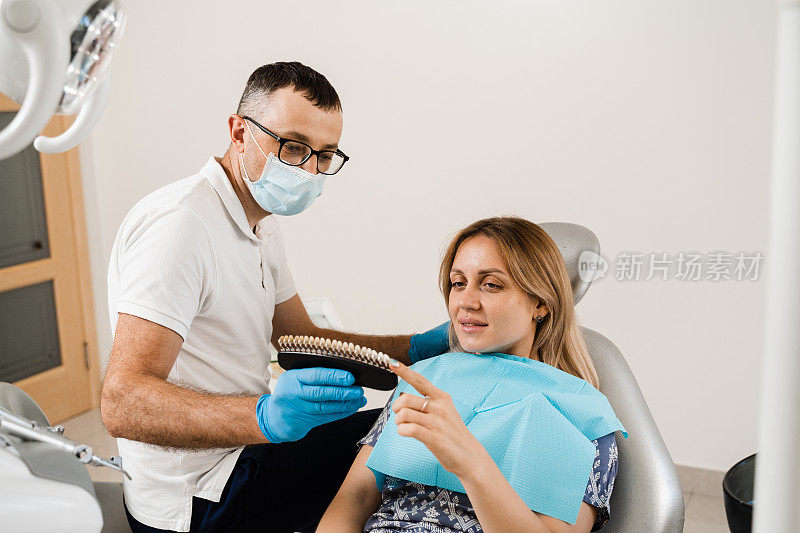牙科医生在牙科诊所为女性病人示范牙齿美白的颜色深浅。牙医。妇女看着贴面或种植牙配色样本在医生手中。