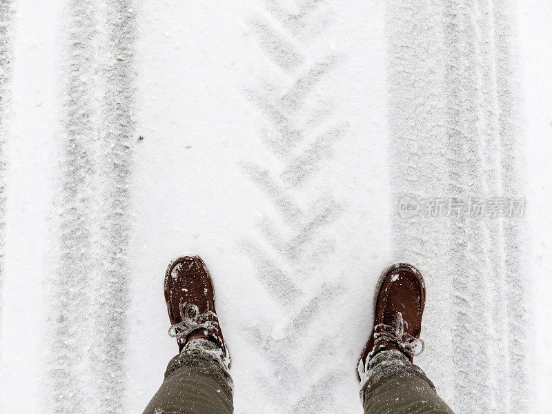 一个穿着棕色靴子的男人站在一条被雪和拖拉机轮胎印覆盖的路上。