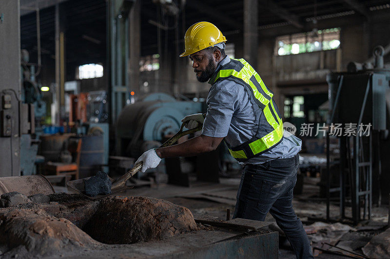 非洲裔美国男性工人穿着背心和头盔安全使用铁锹在金属工厂的地面上挖煤燃烧。在工业工厂操作重金属机械的技工领班。
