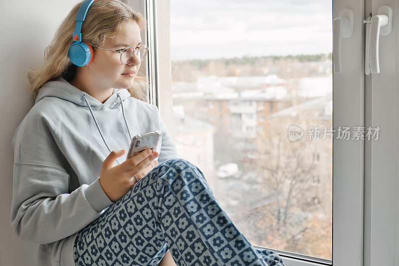 美丽的年轻女孩戴着眼镜，手里拿着智能手机，坐在窗台上，戴着耳机听音乐。