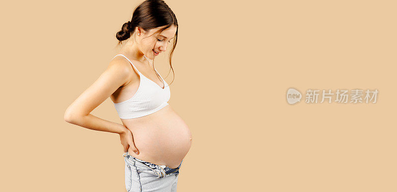 在米黄色的背景上，一个快乐而自豪的孕妇穿着白色上衣和蓝色牛仔裤，看着自己的肚子。
