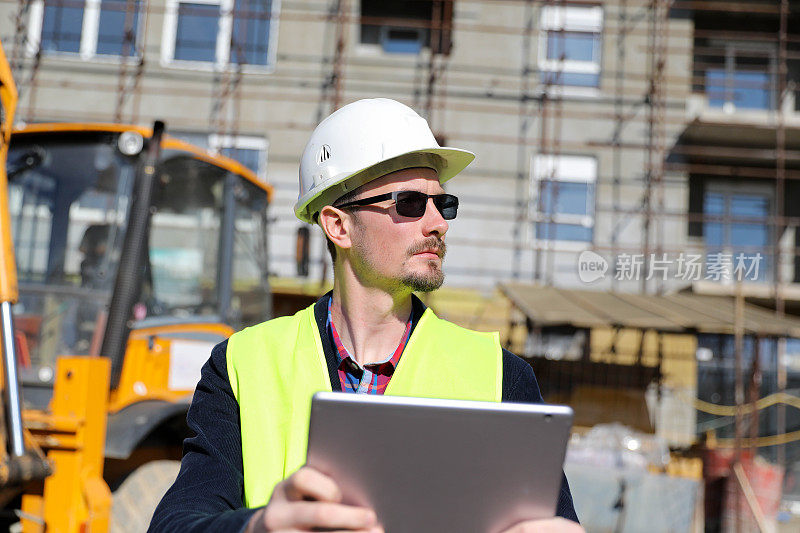 男建筑师在工地使用数码平板电脑，他穿着防护背心和头盔