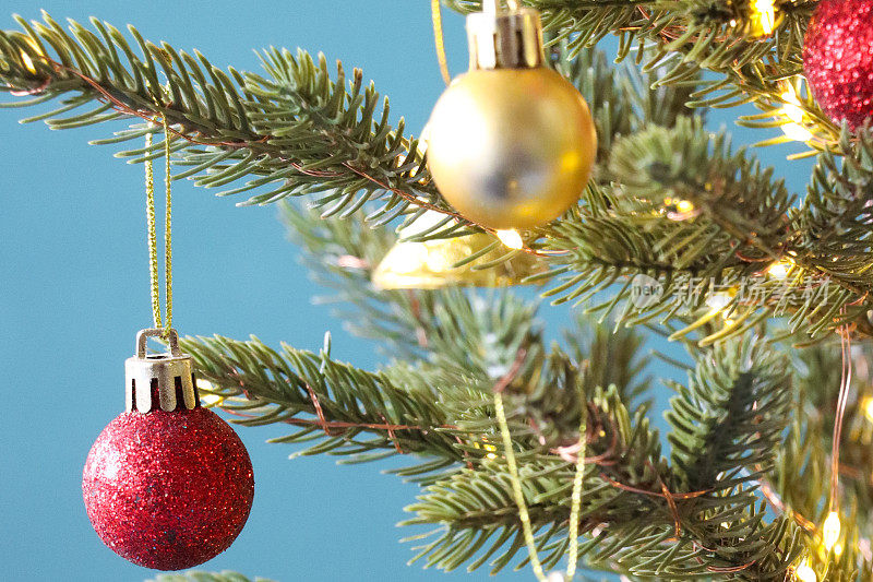 特写:圣诞树上的金线上挂着红色的闪光和金色的小玩意，绿色的云杉针，模糊的白色仙女灯，蓝绿色的背景