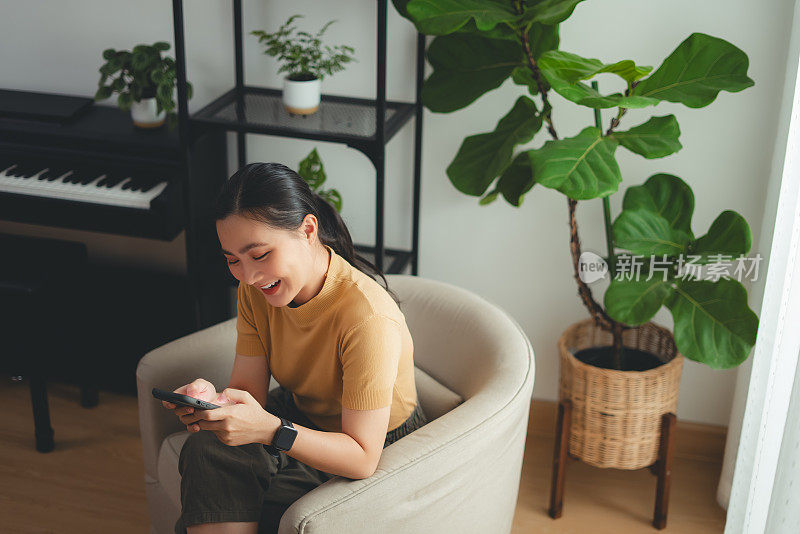 亚洲女人开心地微笑着坐在扶手椅上，在家用智能手机。