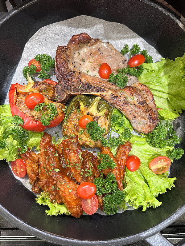 一个金属烹饪锅的特写，上面铺着防油的羊皮纸，里面放着中国猪排、四川鸡、半红半绿的甜椒、生菜叶沙拉、圣女果和欧芹