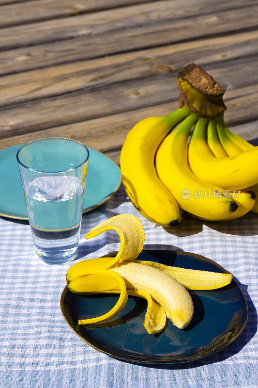 一串熟了的黄香蕉，盘子里放着一根香蕉，蓝色桌布上放着一杯水。阳光明媚的日子里，木质表面上的有机水果。静物照片。有选择性的重点。模糊的背景。