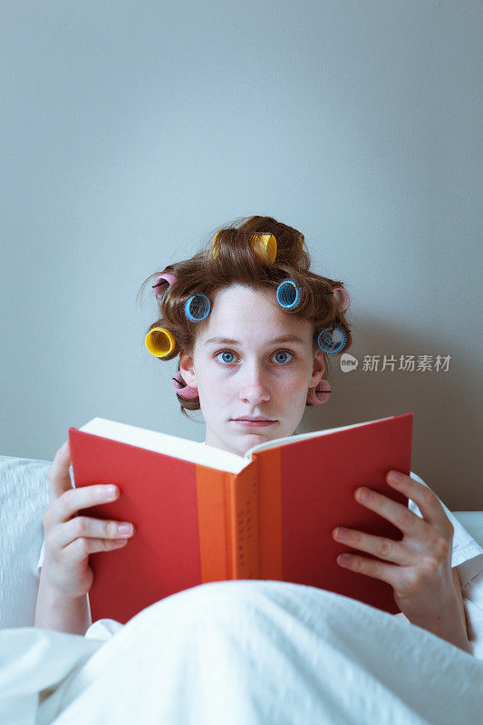 拿着卷发器的年轻女子正在床上看书