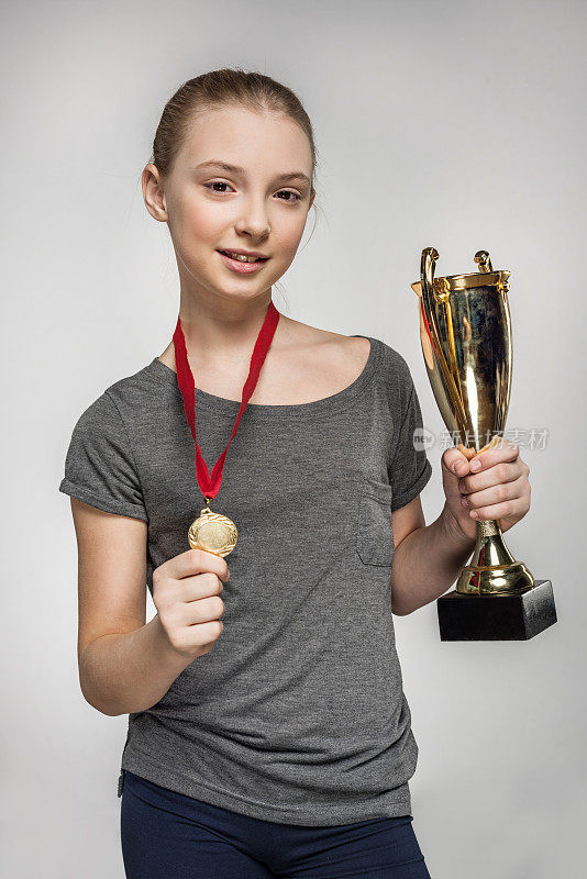 微笑的女孩在运动服拿着奖杯和奖牌孤立在灰色